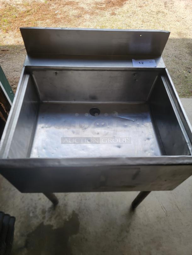 Stainless steel industrial sink with high backsplash, visible wear. Auction tag with the number 12 on the top right corner.
