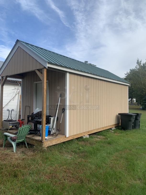 Tan storage shed with green metal roof, porch area, and various garden tools. Good condition, wooden structure, no visible tags.