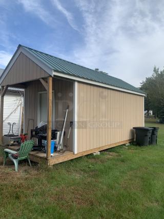 Tan storage shed with green metal roof, porch area, and various garden tools. Good condition, wooden structure, no visible tags.
