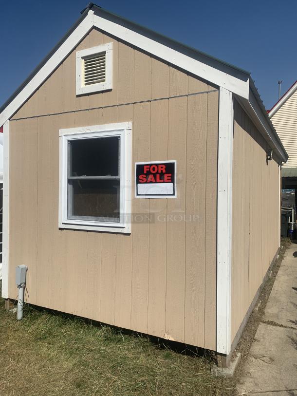 Small wooden shed for sale, beige with white trim, single window, vented top, "For Sale" sign attached.