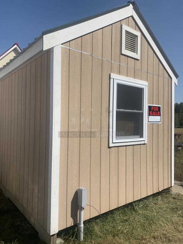 Tan storage shed with white trim, vent and window, "For Sale" sign displayed. Appears in good condition.