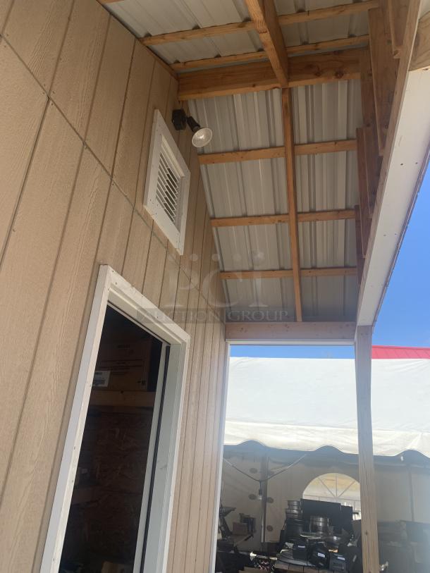 Outdoor wooden shed with vent, exterior light, and white framed door. Seen partially under a metal roof. Excellent condition.