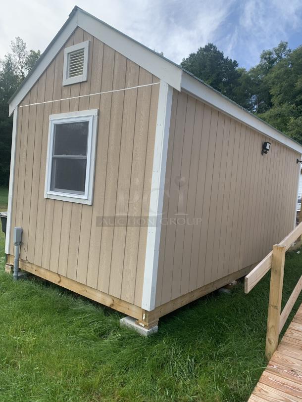Tan wooden storage shed with white trim, single window, roof vent, and mounted floodlight. Sturdy condition on grassy base.