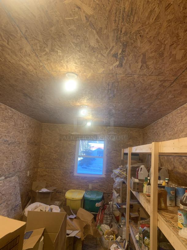 Wooden storage room with shelves holding various items, including bottles and cans. Boxes and bins are on the floor. Room has a window and ceiling lights. Walls are unfinished OSB wood panels.