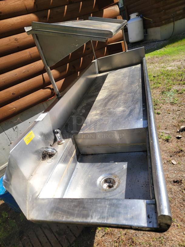 Stainless steel outdoor sink with two compartments, faucet, and drainage. Features shelves and a yellow safety label. Used condition.