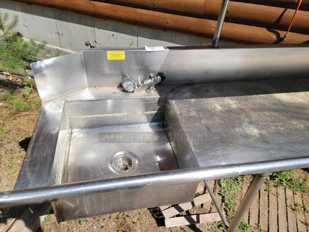 Stainless steel outdoor sink with faucet and pressure gauge; surface shows signs of wear. Safety warning label visible.