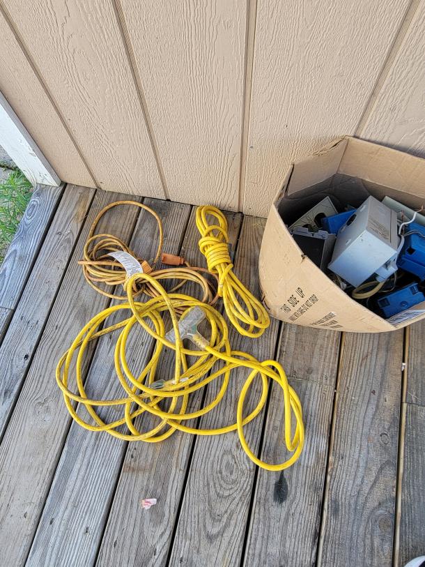 Box of assorted electrical supplies and yellow extension cords on a wooden deck, ideal for wiring projects.