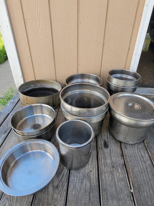 Assorted stainless steel pots and containers on wooden surface, ideal for camping and outdoor cooking; varying sizes and some with lids.