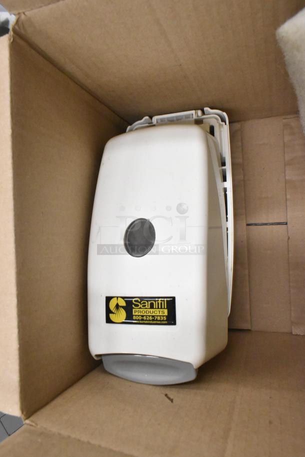 A Sanifil dispenser in a cardboard box, white with a gray button. Label shows "Sanifil Products" and contact information.