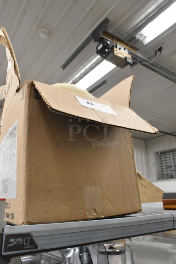 Cardboard box containing a vacuum head, round cleaning pad, and dispenser on a metal shelf. Box shows signs of wear with a label marked "44."
