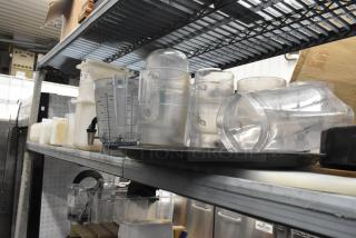 Lot of clear poly pitchers, bins, and dispenser on metal shelving, showing measurement markings and varied sizes, in used condition.
