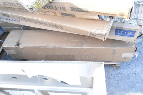 Stack of boxes containing scratch and dent items including wire shelves and bi-fold tables, boxes show 24"x60" dimensions.