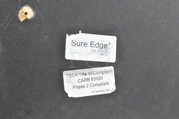 Round tabletop with "Sure Edge" label and compliance markings. P/N: 2010320, TSCA Title VI, CARB Phase 2 compliant.
