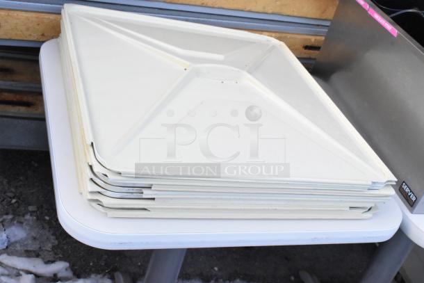Stacked white poly lids on a white dining-height table, next to metal baking pans. Auction lot includes three tables.