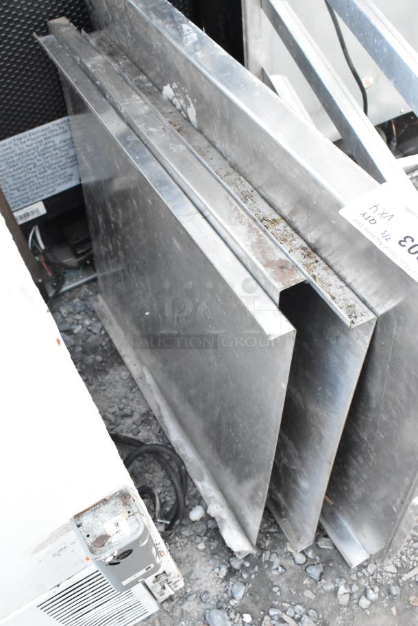 Four metal panels stacked, showing signs of wear and slight rust, positioned on a gravel surface. A label reads "4X."