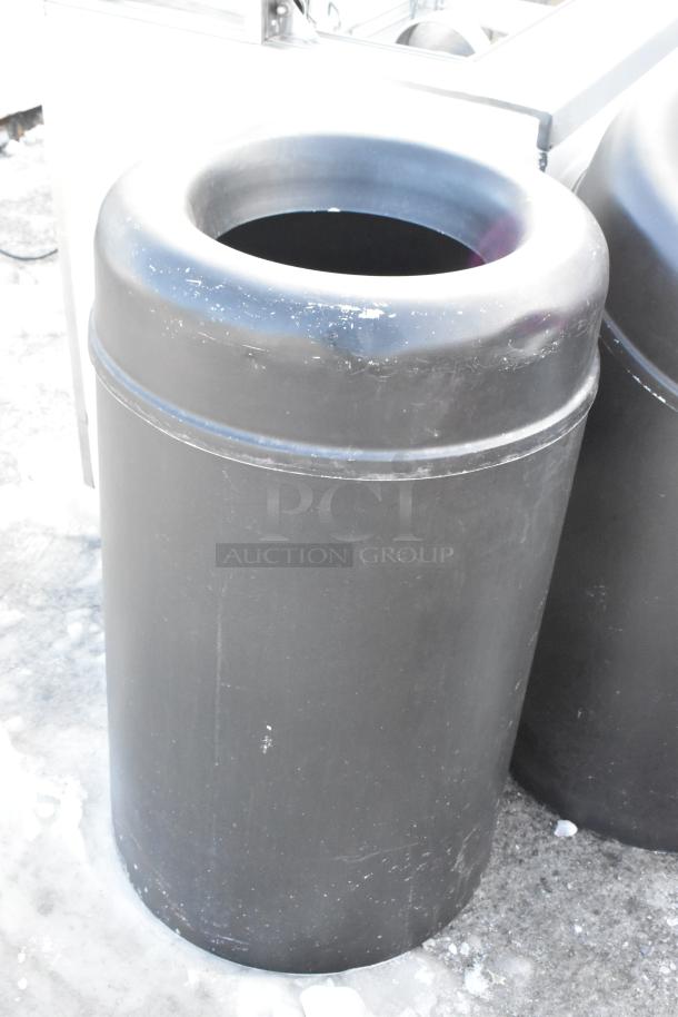 Black trash can, cylindrical shape, plastic material, displays wear. Part of a set with a wooden shell and another black trash can.