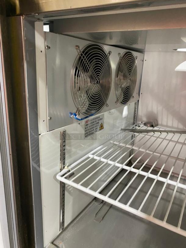 Commercial refrigerator interior with dual fans, wire shelves, and stainless steel finish; label displays safety and operational info.