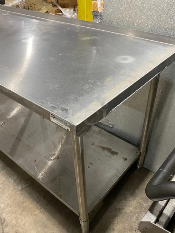 Stainless steel work table with bottom shelf, used condition, visible surface scratches and labels indicating brand and size.