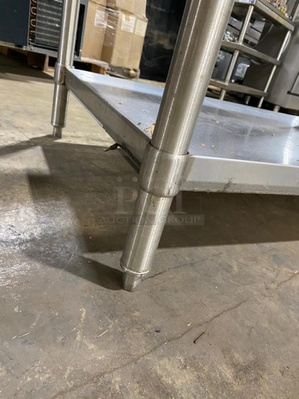 Stainless steel commercial table, sturdy legs, lower shelf, used condition, industrial setting; ideal for kitchen or storage use.