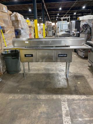 Stainless steel commercial sink with dual basins labeled "Wash" and "Sanitize," featuring drainboard, in used condition.