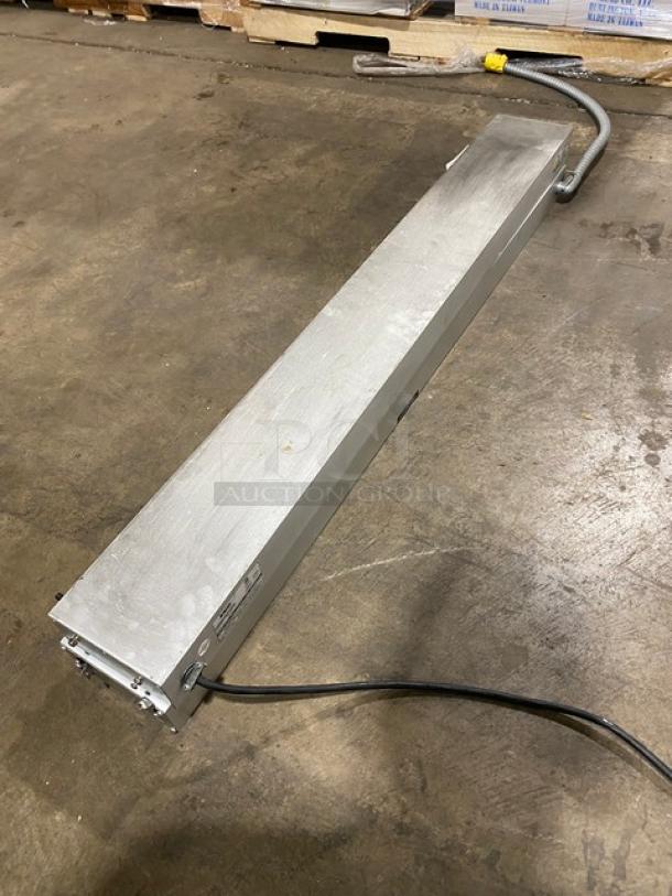 Silver metal industrial equipment box with attached cables on concrete floor. Features power input/output labels and model markings.