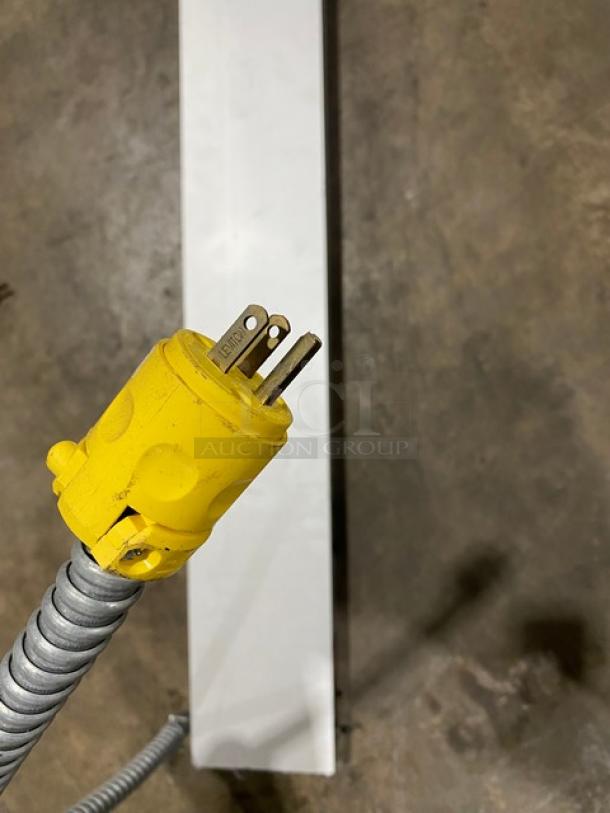 Yellow electrical plug with three prongs on a flexible metal conduit, shown against a concrete floor.