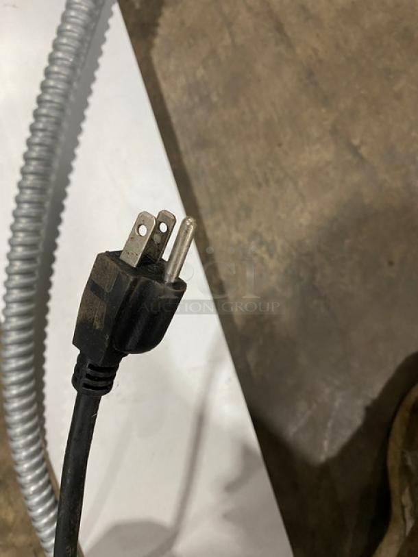 Black three-prong power plug with visible wear on a concrete surface, attached to a coiled, silver conduit.