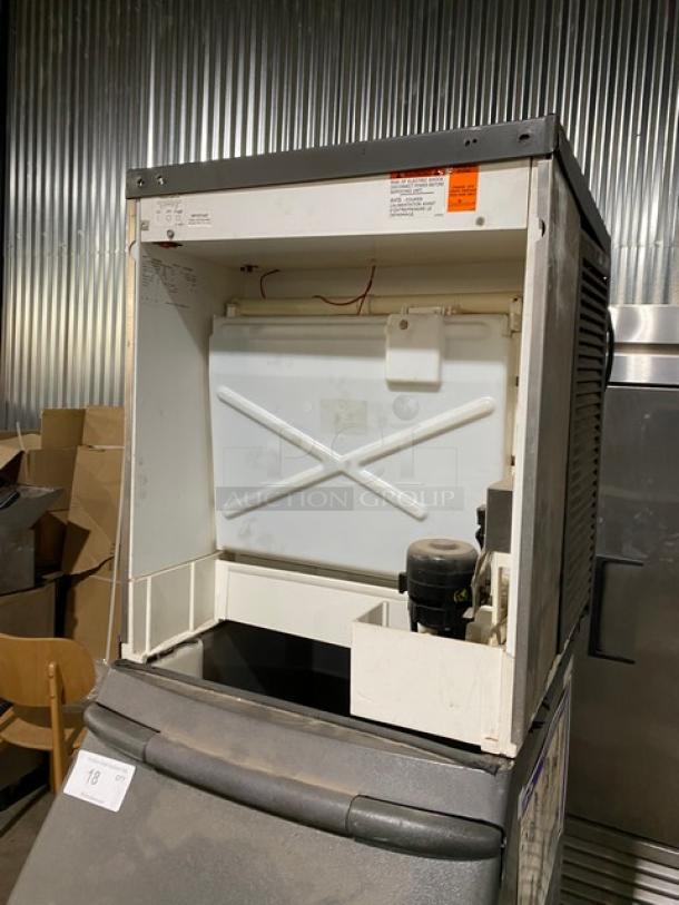Industrial ice machine in used condition, visible internal components and wiring, metal housing with minor wear. Exterior labels include model and safety information.