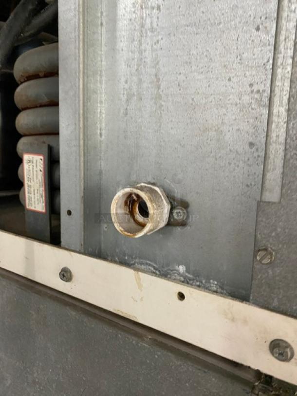 Metal component with visible fitting and wear, mounted on machinery. Label nearby includes instructions and maintenance warnings.