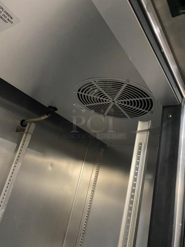 Interior view of a commercial refrigerator showing cooling fan and shelving brackets. Features stainless steel surfaces. Tags are present but not legible.