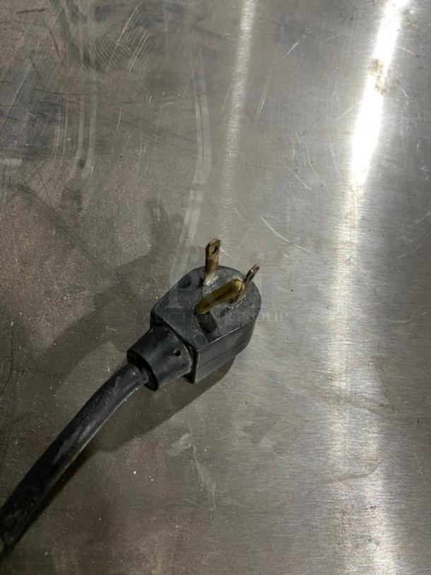 Black three-prong electrical plug on a metal surface, in used condition.