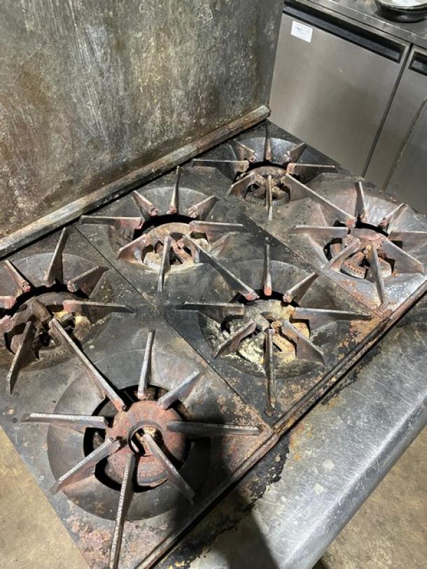 Commercial six-burner gas stove, well-used condition, visible rust and wear, sturdy cast iron grates, ideal for heavy-duty cooking.