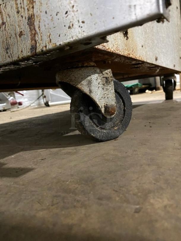 Rusty metal caster wheel attached to a worn industrial cart. Visible rust and wear indicate heavy use and age.
