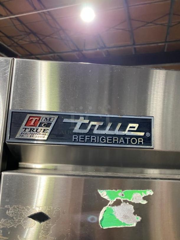 True refrigerator, stainless steel, visible logo plate. Minor surface wear with stickers, indicating used condition.
