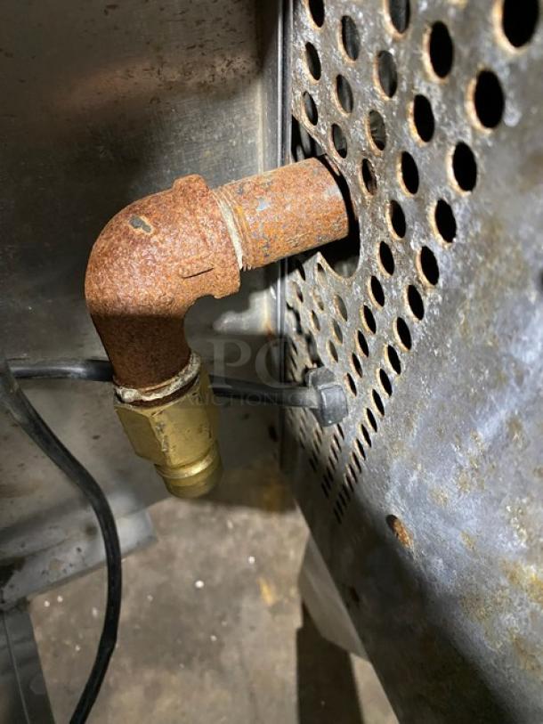 Rusted metal pipe with elbow joint attached to perforated metal panel, showing signs of oxidation.