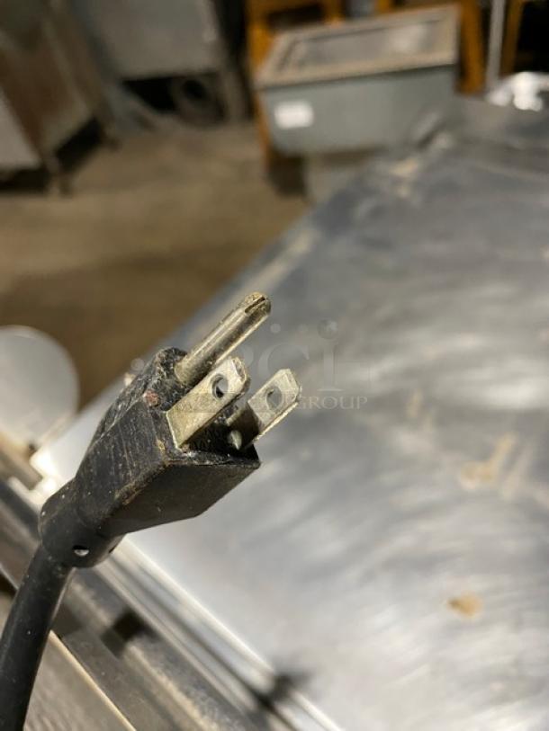 Close-up of a worn electrical plug with three prongs, showing signs of use or damage, against a metal surface background.