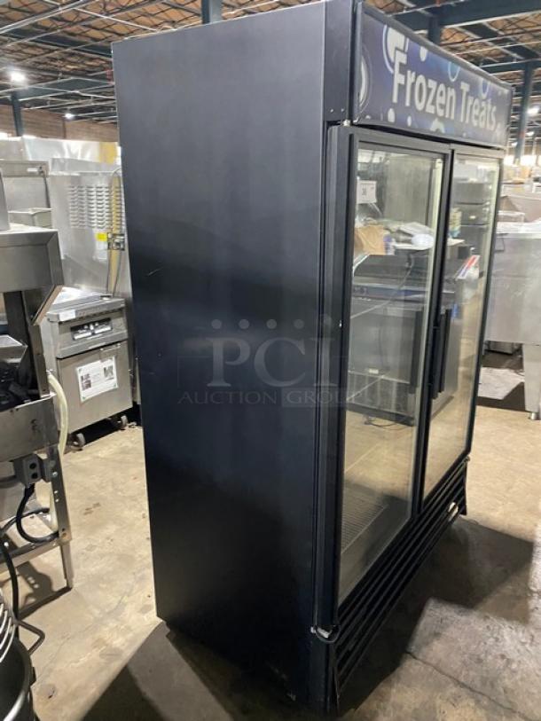 Commercial glass door freezer, two-door, "Frozen Treats" sign, black exterior, used condition, suitable for storing frozen goods.