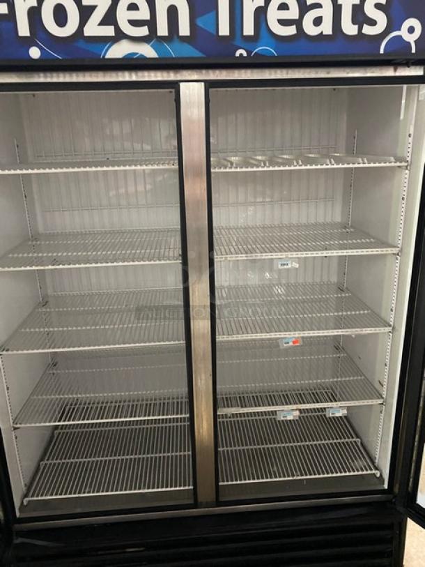 Commercial glass-door freezer with double doors, multiple wire shelves, visible wear, suitable for frozen goods display.