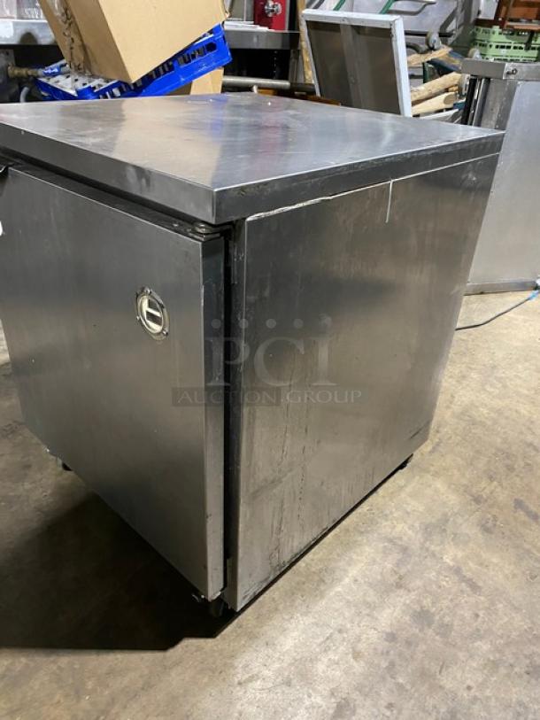 Stainless steel commercial refrigerator, single-door, used condition, visible scratches, lockable handle, industrial setting.