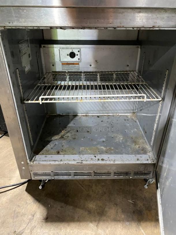 Commercial stainless steel refrigerator with wire shelf, visible wear and rust, adjustable thermostat, on caster wheels.