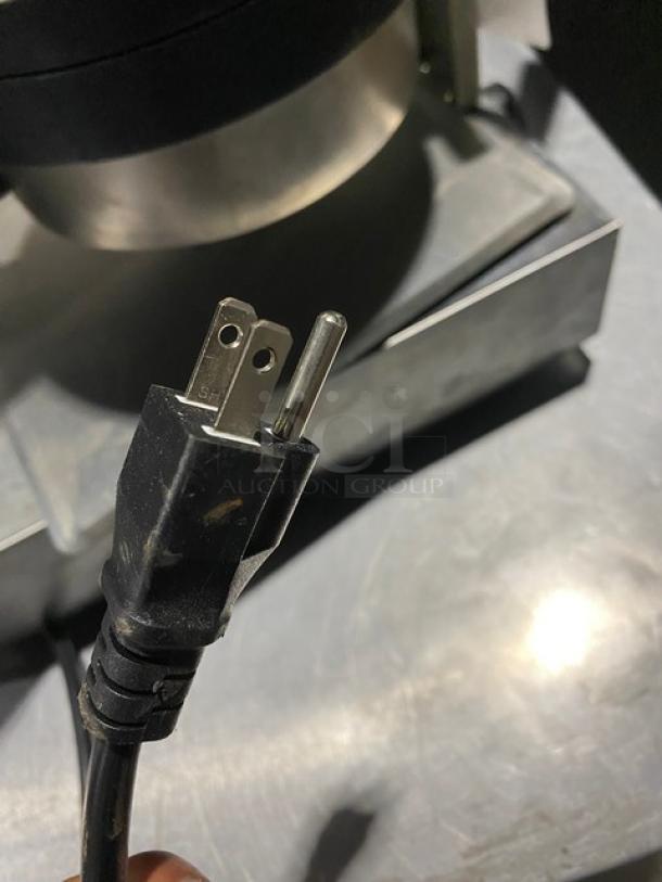 Three-prong electrical plug, black with slight wear, attached to a cable. Black and silver appliance visible in the background.