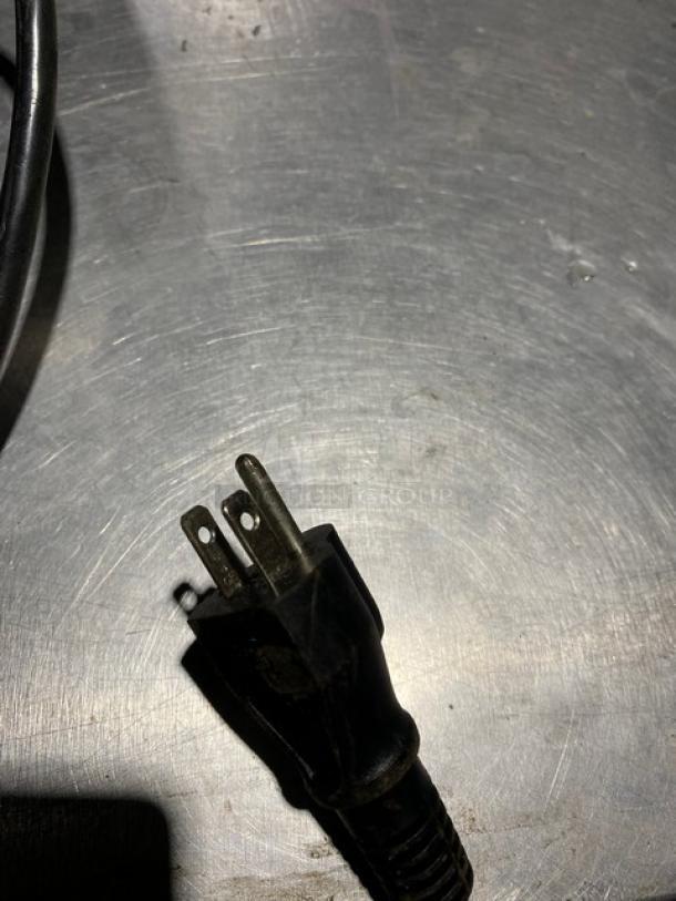 Black power cord plug with three prongs, shown on a metallic surface. Slight wear visible. No markings present.