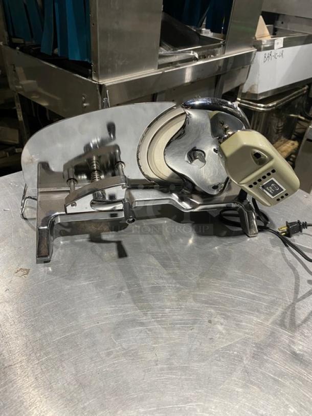 Electric deli slicer on a stainless steel table, featuring a metal blade, adjustable thickness dial, and a power cord.
