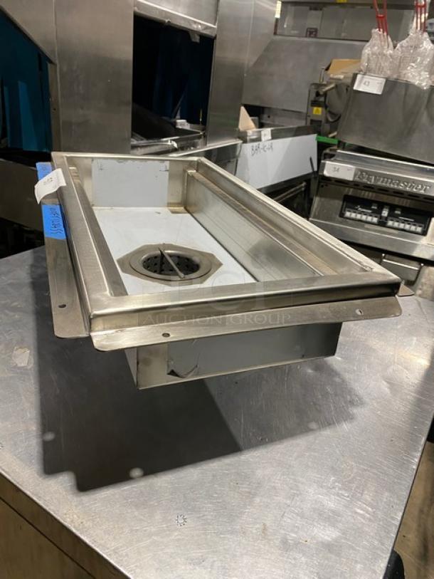 Stainless steel floor sink with drain, used condition, equipped with mounting flanges. Visible tag reads model and size details.