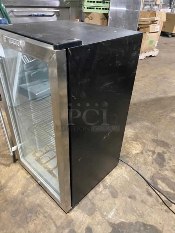 Commercial glass-front refrigerator, black and silver, with shelves. Visible wear and power cord. Ideal for retail display.
