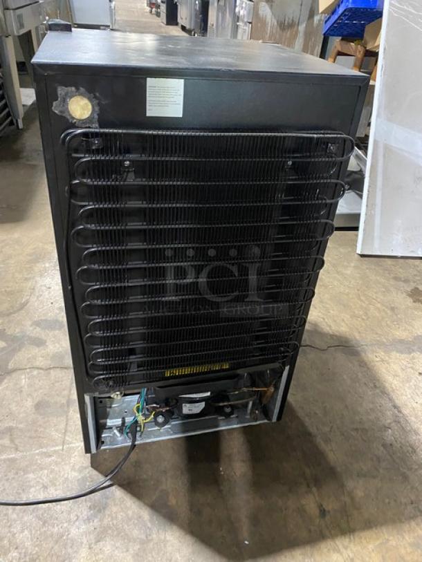 Commercial refrigerator rear view showing black cooling coil, moderate wear. Label indicates model number and maintenance instructions.