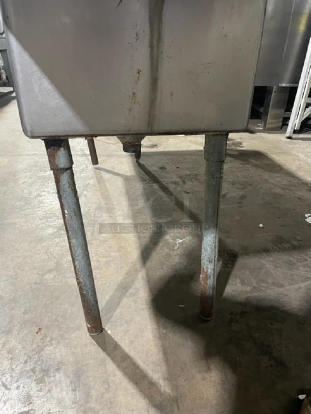 Commercial stainless steel 3-compartment sink with left/right drain boards, backsplash, on legs, signs of rust on pipes.