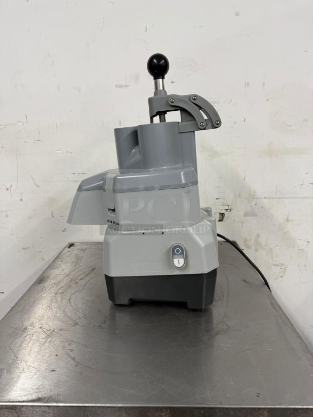 Commercial-grade food processor, gray and black, with manual feed lever. In good condition, power switch visible.