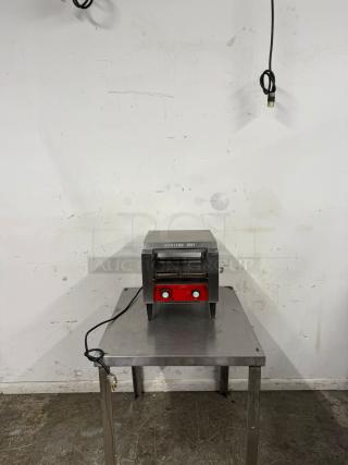 Commercial conveyor toaster on stainless steel table, red controls, labeled "Caution Hot," power cord included.