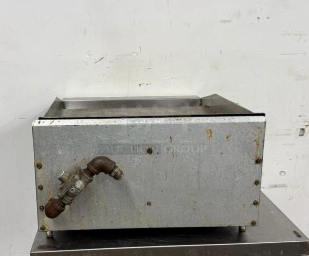 Commercial stainless steel griddle with visible wear and rust spots, featuring a pipe connection on the side.